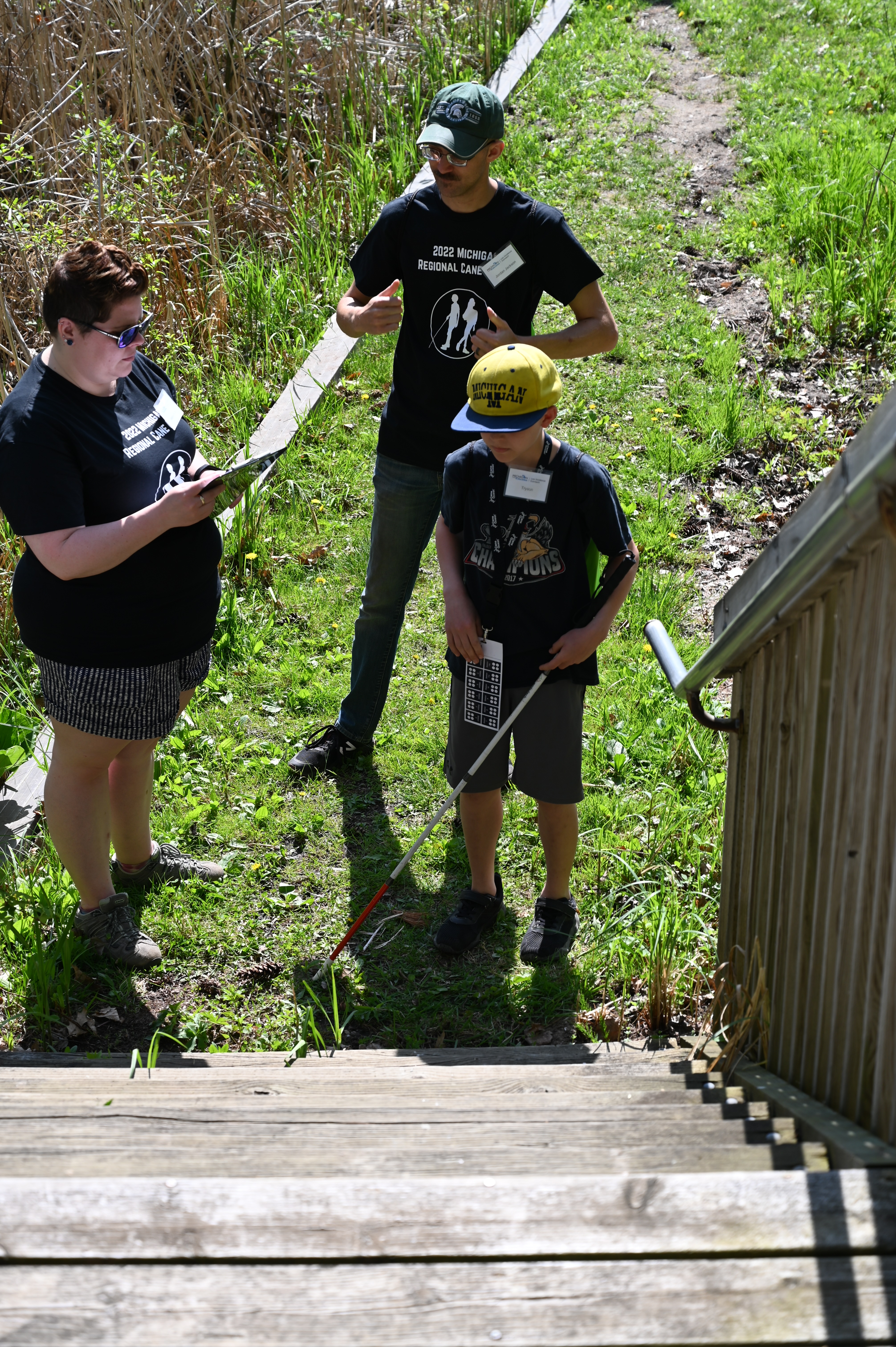 A boy navigates with a cane outside while volunteers give instructions.