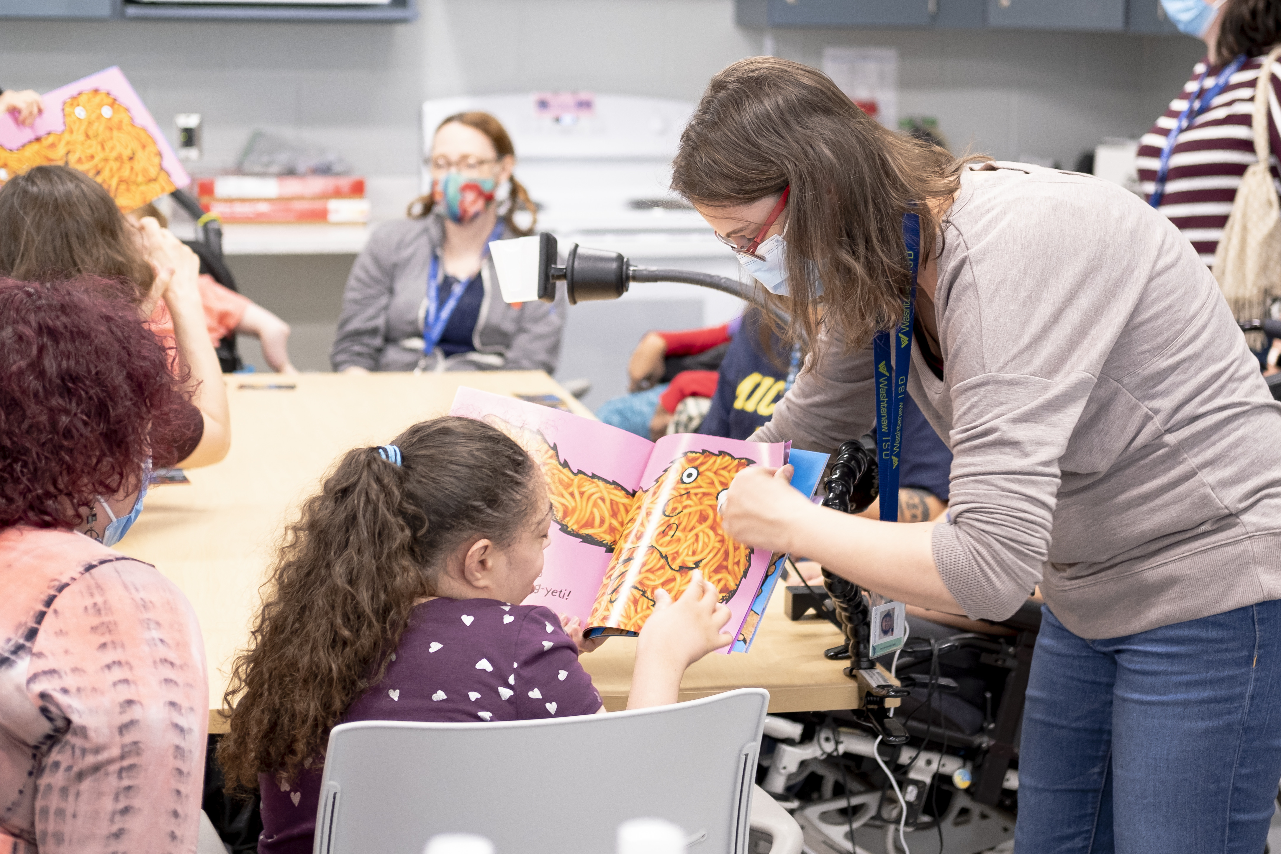 A girl who is sitting at a table full of students and teachers touches a colorful illustrated book that her teacher is holding open.