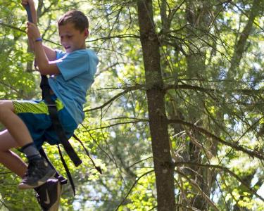 A boy smiles while rappelling down a wooden climbing wall in the woods.