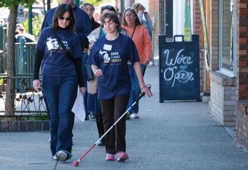 A student and volunteer walk down a busy city street wearing Cane Quest T-Shirts