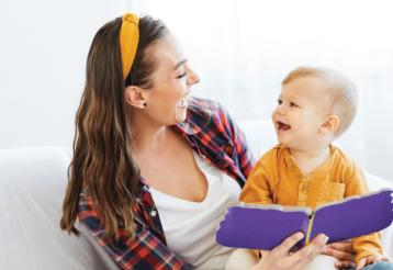 A mother sits on a couch with her toddler in her lap. The both smile as the mother reads a book. 