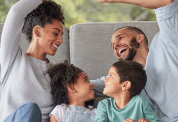 A smiling family sits in front of a couch. Two adults give a high five. 