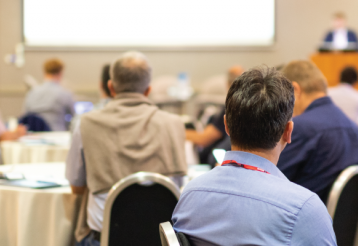 Audience members attentively watching a presentation at a conference, with tables and a speaker in the background.