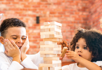 A family plays Jenga at a table, laughing and focused on balancing wooden blocks, with a rustic brick background.
