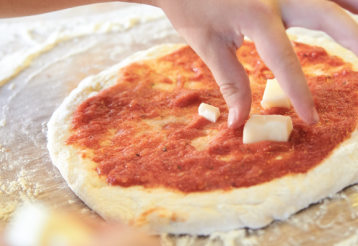 A child's hand adds cubes of cheese to a pizza topped with tomato sauce, on a floured countertop.