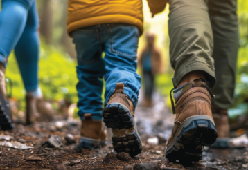 A close-up of feet walking on a forest trail, highlighting a child and two adults in colorful outdoor attire, surrounded by lush greenery.