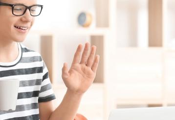 A teenager wearing a hearing device smiles and waves during a video call while holding a mug.
