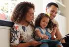 A mother, father, and young daughter smile while reading a book together at home.