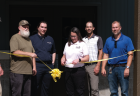 Nine people stand in front of Oak Recreation Center, which has a yellow ribbon draped across it. Jill Teegardin has just cut the ribbon with scissors.