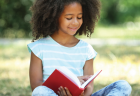 A young girl reading a book outside on a sunny day.