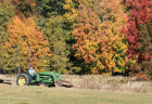 Parents and children ride in a trailer pulled by a tractor in a large field surrounded by trees in fall.