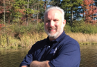 Crossing his arms, Matt Polzin smiles at the camera while standing next to a lake in a wooded area.