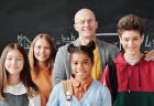 A teacher smiles for a photo with six pre-teen students in front of a chalkboard.