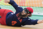 Goalball player John Kusku, wearing an eye cover, slides sideways and stretches his arm out to block a goal.