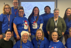 A group of fourteen teacher consultants pose for a group photo in a large room, mostly wearing matching blue shirts.