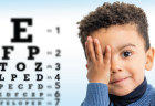 Young child in a blue sweater covering one eye, next to an eye chart during an eye exam.