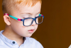 A young child with short hair reads a book while wearing colorful blue and pink glasses.