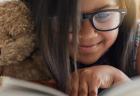 A young girl smiles while holding a teddy bear and reading a book