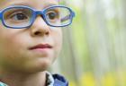 A young boy wearing glasses and a hearing device smiles while standing outside.