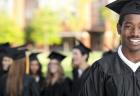 A smiling graduate in a black cap and gown smiles for a photo in front of students who are talking in a group.