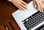 Closeup of hands resting on a silver laptop that is sitting on a brown wooden table.