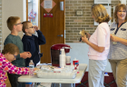 One girl sits and two boys stand at a folding table that features a single-cup coffee machine, cups, and bottles of water. One of the boys talks to a woman who is in line to buy coffee on the other side of the table.