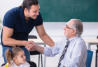 A parent who is standing next to a young girl smiles and shakes hands with a teacher who is sitting in a chair in his classroom.