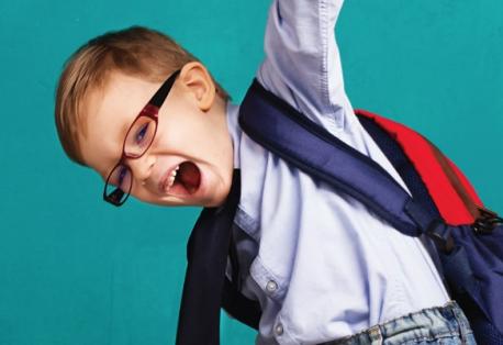 A young boy wearing glasses and a backpack smiles while raising one arm in the air.