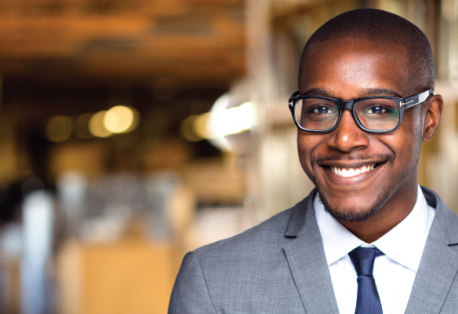 A man wearing a suit and glasses smiles at the camera while standing in a library.