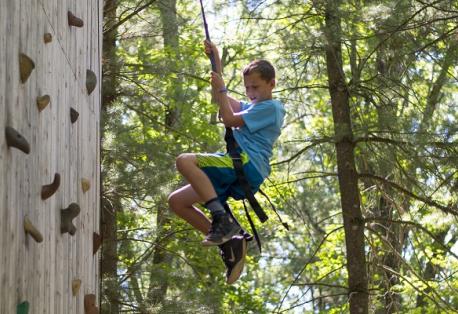A boy smiles while rappelling down a wooden climbing wall in the woods.