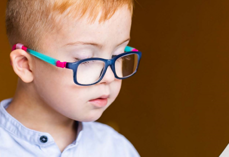 A young child with short hair reads a book while wearing colorful blue and pink glasses.