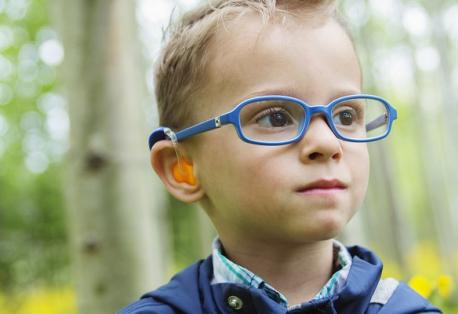 A boy wearing glasses and a hearing device smiles off-camera while standing in a wooded area.