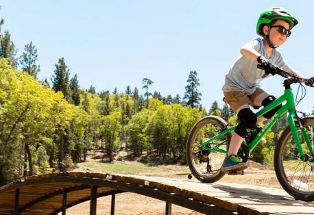 A young boy smiles while riding a bike over a wooden ramp on a sunny day.