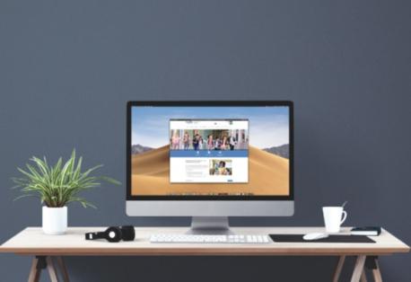 A large computer monitor on a desk, showing the MDE-LIO website. The desk features a plant, headphones, keyboard, and mug.