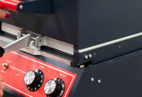 Closeup of a woman's hands pressing the top of a thermoform machine toward the base. A small red instrument panel on the front features a switch and knobs for time and heat.
