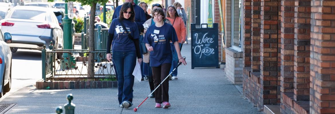 A student and volunteer walk down a busy city street wearing Cane Quest T-Shirts