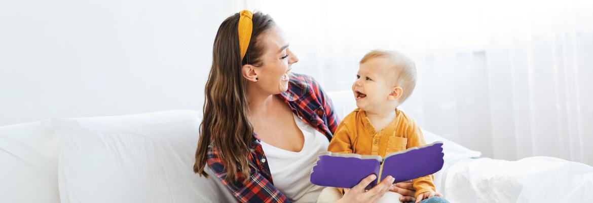A mother sits on a couch with her toddler in her lap. The both smile as the mother reads a book. 