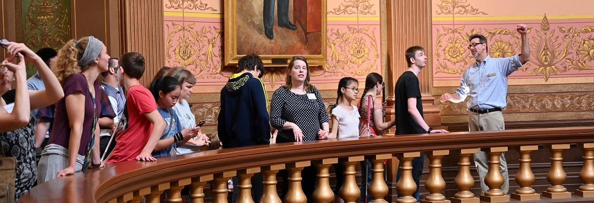 Several adults and children stand next to a large circular railing as a tour guide describes features of the Lansing capitol building.