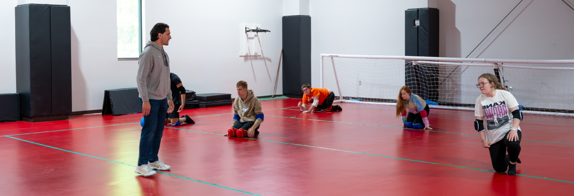 An instructor teaches a group of students how to play goal ball in a gym.