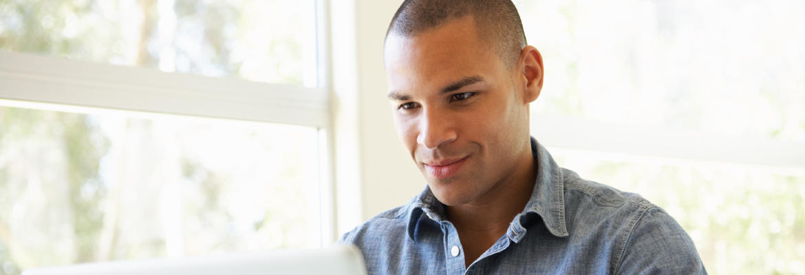 Closeup of a man smiling at a laptop while sitting next to a large window.
