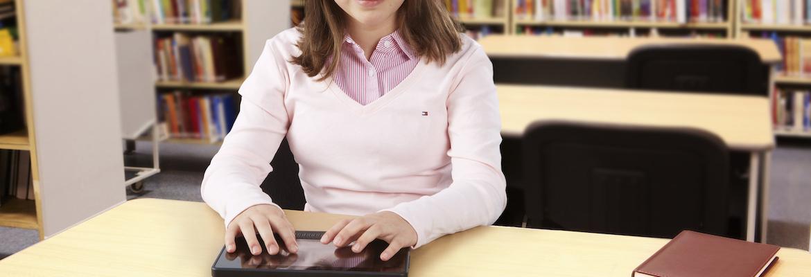 Closeup of a young girl sitting at a table in a library and placing her hands on a BrailleNote Touch.