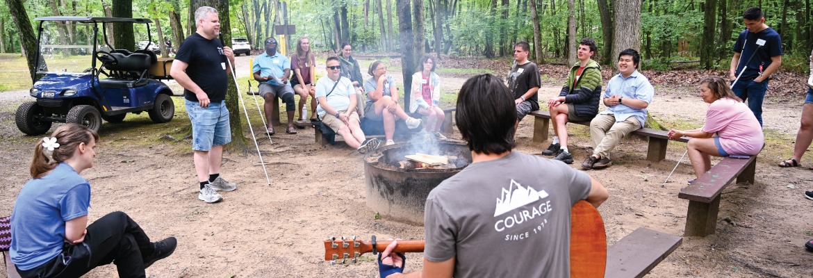 A group of people gathered around a campfire, enjoying music and conversation in a wooded outdoor setting.