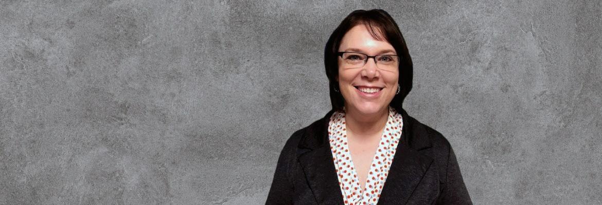 Amanda, wearing glasses, a white dotted shirt, and a black blazer, smiles for a photo in front of a gray background.