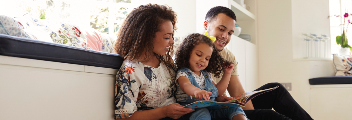 A mother, father, and young daughter smile while reading a book together at home.