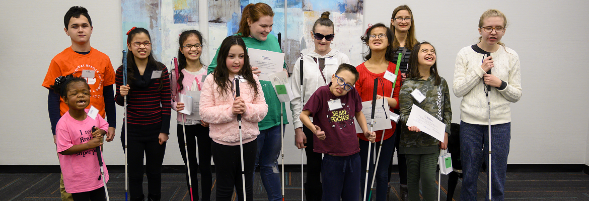 Twelve students, ranging from elementary to high school age, smile for a group photo. Most of the students are holding white canes.
