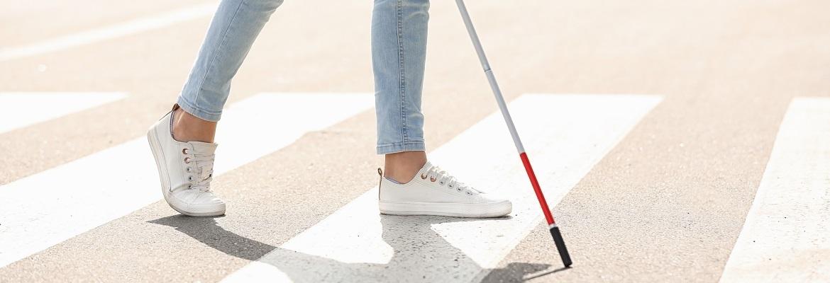 Closeup of someone's legs and shoes as they navigate a crosswalk with a white cane.