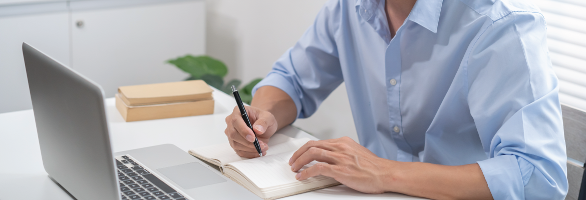 Person writing in a notebook at a desk with a laptop and stacked books in a bright, minimal office setting.