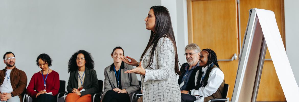 A speaker presents in front of an audience seated in a meeting room, engaged in discussion and learning. A whiteboard is visible.