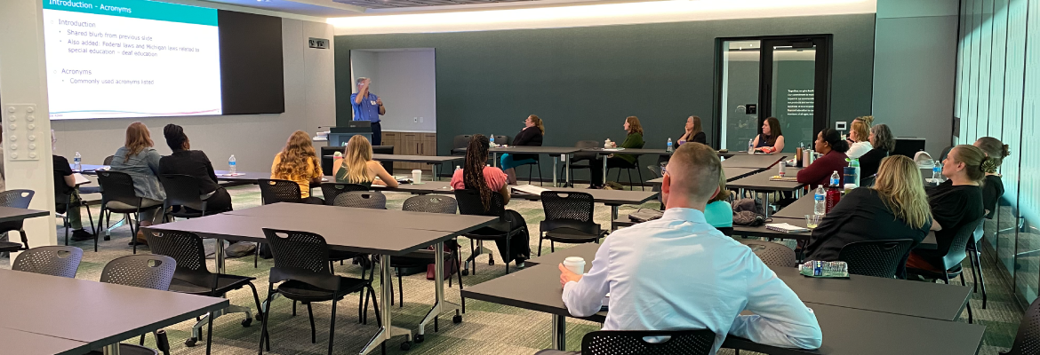 Classroom setting with attendees seated at tables listening to a presenter.