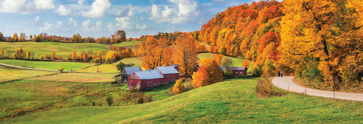 A group of red farm buildings located on rolling hills bordered by trees with yellow and orange leaves. 